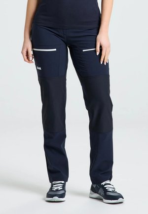 El modelo lleva pantalones utilitarios azul marino con bolsillos de cremallera blancos y zapatillas deportivas negras y blancas, sobre un fondo claro de estudio.
