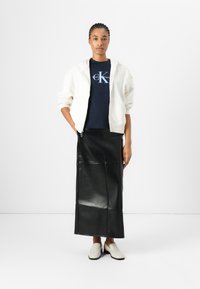 Felpa bianca con zip sopra una T-shirt blu navy di Calvin Klein, abbinata a una lunga gonna di pelle nera e scarpe slip-on beige.