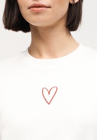 Γυναίκα με κοντά σκούρα μαλλιά φορώντας λευκό πουκάμισο με απλό κόκκινο περίγραμμα καρδιάς στο ύψος του στήθους.