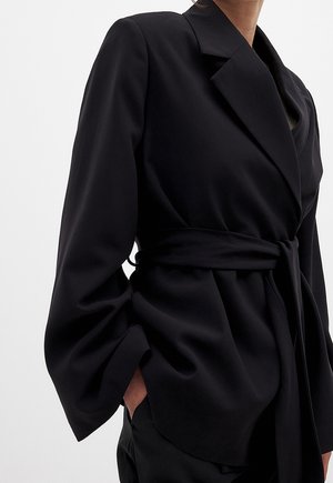 Frau trägt einen schwarzen Blazer mit Gürtel, breiten Revers und langen Ärmeln, Hand teilweise in der Tasche, dargestellt vom Hals bis zu den Hüften.