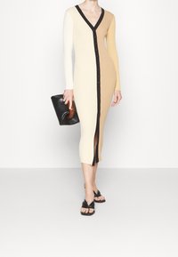 Abito a maglia a coste in beige e crema con maniche lunghe, scollo a V, chiusura frontale con bottoni e spacco laterale; abbinato a una pochette nera e sandali.