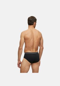 Uomo con capelli corti e scuri, visto di spalle, indossa slip neri con fascia in vita a righe bianche su sfondo semplice.