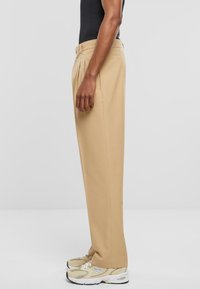 Beige plissierte Hose mit geradem Bein, aus einem glatten Stoff gefertigt. Getragen mit einem eng anliegenden schwarzen Oberteil und beigen Sneakers.