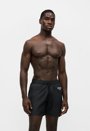 Homme musclé à la peau foncée portant un short de bain noir, debout devant un fond blanc uni.