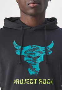 Svart hoodie med en turkos tjurskalle-grafik och "PROJECT ROCK"-text i klargul, med en mjuk yta och justerbar dragsko. 