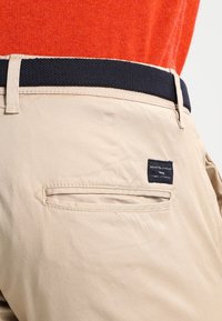 Beiga byxor med en marinblå midja, bakficka och en etikett som säger "SELECTI HOME." Slät tyg med subtila sömnadsdetaljer.