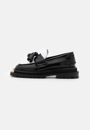 Mocassin en cuir noir avec pampille, semelle épaisse à crampons et détail en métal doré sur l'avant, vu de côté sur un fond blanc uni.