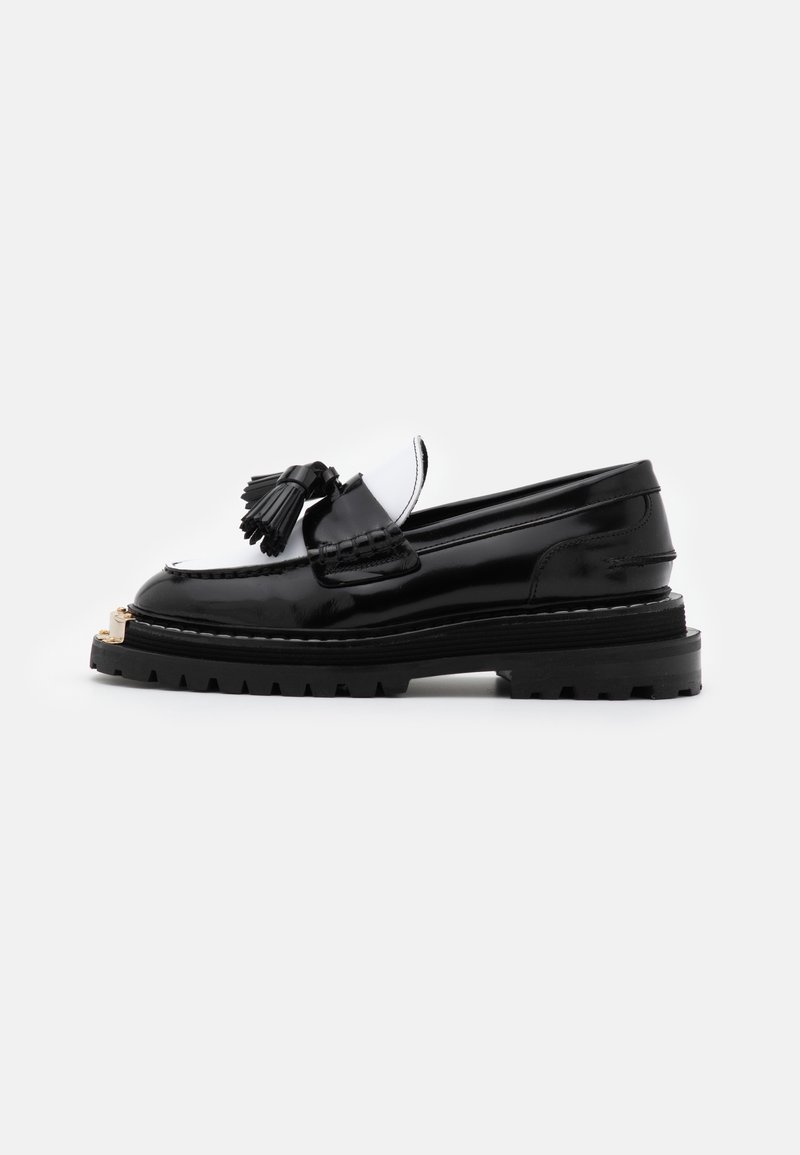 Mocassin en cuir noir avec pampille, semelle épaisse à crampons et détail en métal doré sur l'avant, vu de côté sur un fond blanc uni.