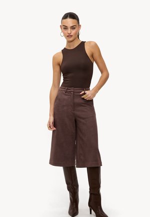 Bruine mouwloze bodysuit gecombineerd met hoge taille, bruine suède culottes en kniehoogte bruine laarzen. Model poseert met één hand op de heup.