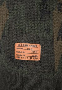 Gestrickter Pullover mit einem Tarnmuster in dunkelgrünen und braunen Tönen. Enthält ein orangefarbenes Stoffetikett mit Produktinformationen.