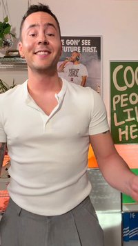 Uśmiechnięty mężczyzna w białej koszuli i szarych spodniach stoi w pomieszczeniu z roślinami i plakatami, w tym jednym za nim przedstawiającym mężczyznę z mikrofonem.