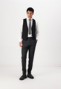 Schwarzweste über einem weißen Hemd, grauer Krawatte und grauen Hosen. Glattes Material, taillierte Passform und Knopfdetails an der Weste. Trägt schwarze Schuhe.