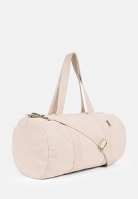 Canvas duffel bag in light beige with dual handles, an adjustable shoulder strap, and a zipper closure. Features a gold-tone hardware accent.