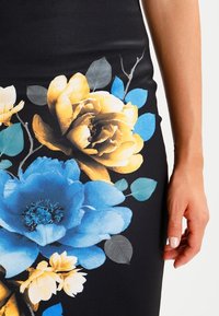 Abito nero con grandi motivi floreali in giallo, blu e bianco, con foglie verdi. Tessuto liscio e aderente.