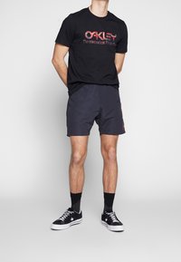 T-shirt noire avec texte graphique rouge, associé à un short sombre. Les chaussures sont des baskets noires avec des semelles blanches et des chaussettes noires. Design simple, coupe décontractée.