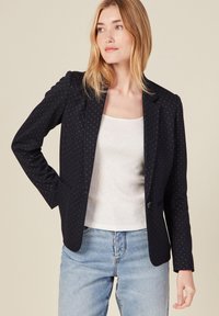 Blazer negro con una textura de patrón sutil, cierre de un botón y dos bolsillos, combinado con una camiseta sin mangas blanca y unos vaqueros azules.