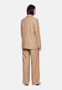 Beige Wollmischung-Anzug mit einem langen Blazer mit Reverskragen und Schlitzen im Rücken, kombiniert mit weit geschnittenen Hosen. Sanfte Textur, minimale Details.