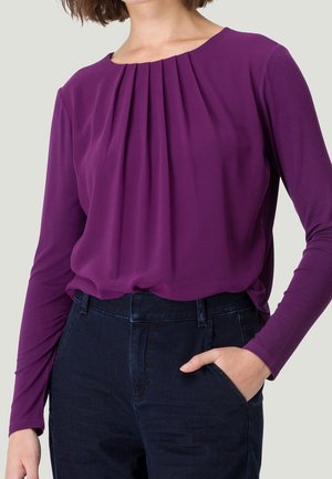 Mujer con blusa morada de manga larga con pliegues delanteros metida en vaqueros azul oscuro de talle alto, una mano en el bolsillo, fondo neutro.