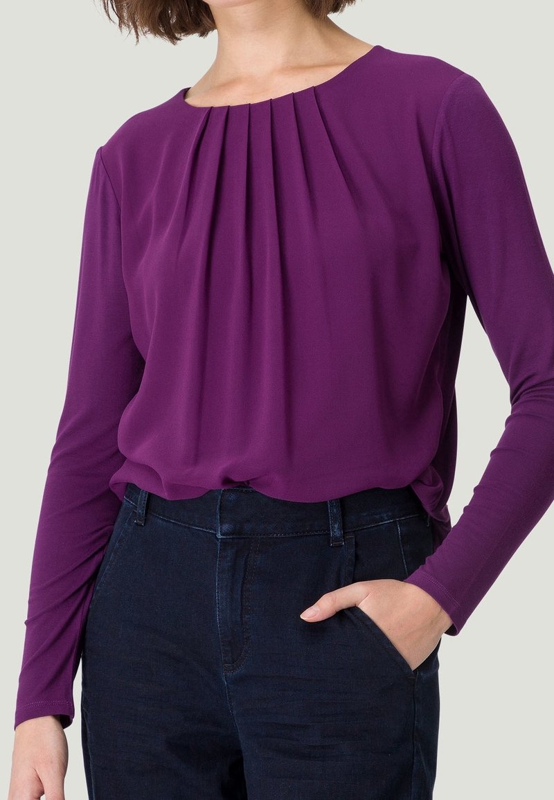Femme portant un chemisier violet à manches longues avec des plis à l'avant, rentré dans un jean taille haute bleu foncé, une main dans la poche, fond neutre.
