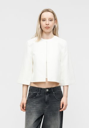 Junge Frau mit glatten blonden Haaren, die eine weiße, kurz geschnittene Jacke mit weiten Ärmeln und ausgewaschene schwarze High-Waist-Jeans trägt.