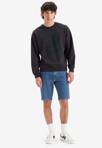 Hanorac gri închis cu text grafic verde care spune "CU TOȚII SUNTEM PRAF DE STELE". Asortat cu pantaloni scurți din denim albastru și adidași albi.