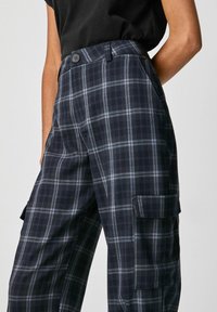 Osoba nosząca ciemne, kratkowane spodnie cargo z bocznymi kieszeniami oraz czarną bluzkę włożoną w spodnie, przedstawiona od pasa do połowy uda.