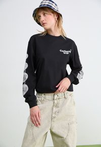 Svart langermet t-skjorte med hvit Carhartt WIP-logo, med grafiske trykk på ermene; kombineres med lyse, baggy denimbukser.
