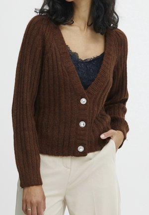 Femme portant un cardigan en maille côtelée marron avec trois boutons décoratifs sur un haut en dentelle noire et un pantalon crème, main dans la poche.