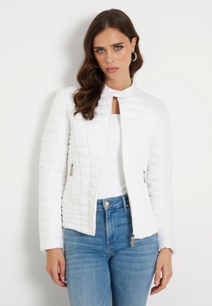 Veste matelassée blanche avec col montant, zips latéraux et silhouette ajustée, associée à un jean léger bleu et un haut blanc.