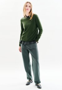 Dunkelgrüner Wollpullover, navy-blaues kariertes Hemd darunter, graue lässige Hose. Schwarze Slipper mit goldenen Akzenten. Glatte Texturen, geschichteter Look.