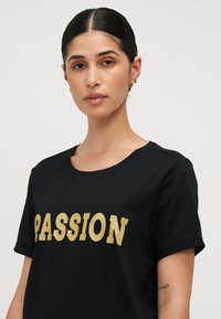 Juodas T-shirt, pagamintas iš minkštos medvilnės, su dideliu auksiniu blizgučiu užrašu "AISTRĄ" fronte ir trumpomis įvyniotomis rankovėmis.