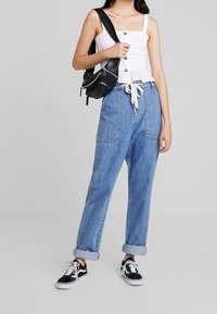 Kobieta w białej bezrękawnikowej bluzce, niebieskich jeansach z wysokim stanem i podwiniętymi nogawkami, czarnych trampkach oraz czarnej torbie na ramię na szarym tle.