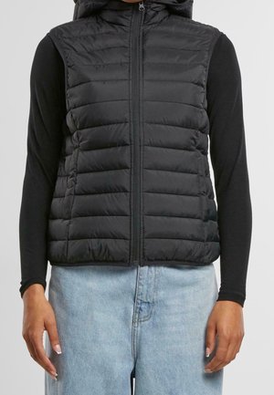 Chaleco negro acolchado sin mangas con capucha, llevado sobre una camiseta negra de manga larga y vaqueros vaqueros azul claro, mostrado desde la mitad del torso hasta las manos.
