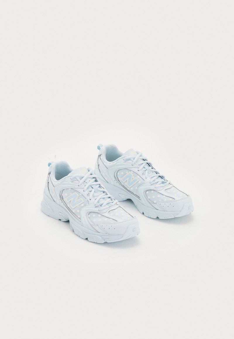 Paire de baskets blanches New Balance avec des touches bleu clair et une fermeture à lacets, présentées sur fond blanc.