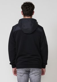 Sudadera negra con textura acanalada y una capucha suave y aislante. Presenta un bolsillo con cremallera en la manga y un corte relajado.