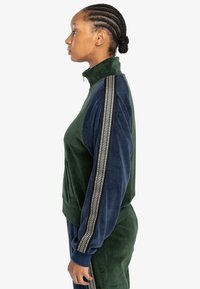 Chaqueta de velour verde y marino con cremallera y cuello alto, que presenta un detalle de raya texturizada en las mangas y un bolsillo frontal.