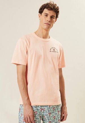 Joven vestido con una camiseta rosa clara que lleva el texto "Off to the Sea" y pantalones cortos azul claro con un patrón de langostas rojas y olas.