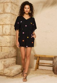 Schwarzer kurzer Overall mit bunter Sonnenstrahlen-Stickerei, lockeren Ärmeln und elastischem Bund. Kombiniert mit schwarzen Riemchensandalen.