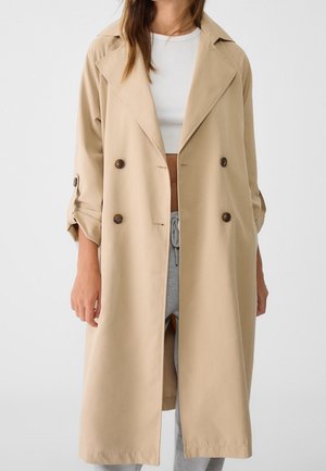 Vrouw draagt beige dubbelgeknoopt trenchcoat over een witte crop top en grijze broek met trekkoord, staand tegen een effen achtergrond.