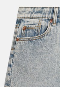 Unos vaqueros de mezclilla de color azul claro presentan un gran bolsillo frontal, remaches de cobre y costuras en contraste con una textura ligeramente deslavada.