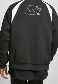 Sudadera negra con paneles laterales blancos, puños y dobladillo acanalados. Presenta un logo bordado en blanco en la parte superior de la espalda. Textura suave.