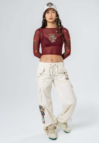 Top de malla burdeos de manga larga con bordado floral, combinado con pantalones cargo crema con bolsillos laterales y patrones coloridos. Una gorra blanca completa el look.