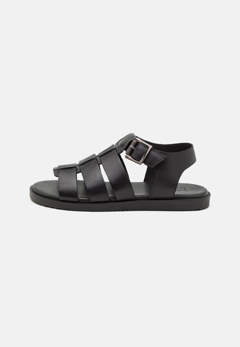 Sandale homme en cuir noir avec trois larges brides, bride de cheville avec boucle métallique et semelle plate sur fond uni.