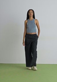 Graues, geripptes Tanktop und schwarze, locker sitzende Cargo-Hosen mit elastischem Bund. Die Schuhe sind hellfarben. Der Hintergrund ist schlicht.