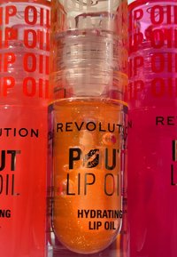 Fuktighetsgivende leppeolje i en gjennomsiktig beholder. Oransje farge med synlig glitring, merket "POUT LIP OIL" med fet svart tekst. Aksenter i rosa og rødt.