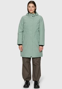 Femme portant un manteau d'hiver à capuche vert clair, un pantalon ample marron foncé et des bottines noires, se tenant devant un fond uni.