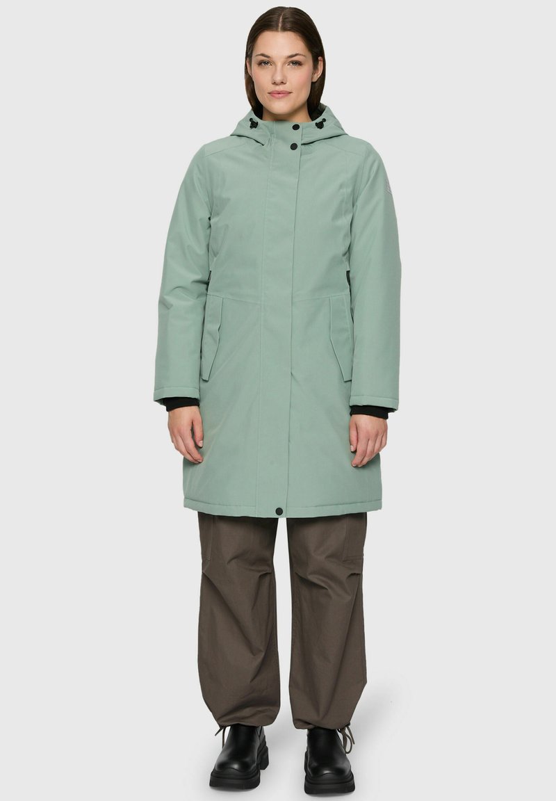 Femme portant un manteau d'hiver à capuche vert clair, un pantalon ample marron foncé et des bottines noires, se tenant devant un fond uni.