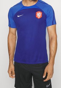 Camisa atlética de color azul royal con mangas cortas, que presenta un diseño texturizado en el hombro azul y un logo de cresta roja. Logo de Nike en blanco en el pecho.