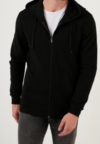 Felpa nera con zip, realizzata in un tessuto texturizzato, dotata di cappuccio con coulisse regolabile e tasche frontali. I polsini e l'orlo a coste completano il design.