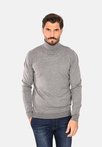Maglione grigio a collo alto in un tessuto morbido con un design aderente. Presenta polsini e vita a costine, indossato sopra jeans denim scuri.